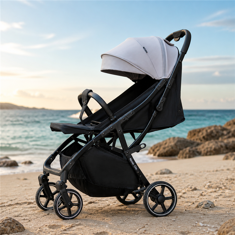 Black-frame baby stroller with gray canopy, reclining seat, four wheels and under-seat storage basket, parked on a sandy beach w